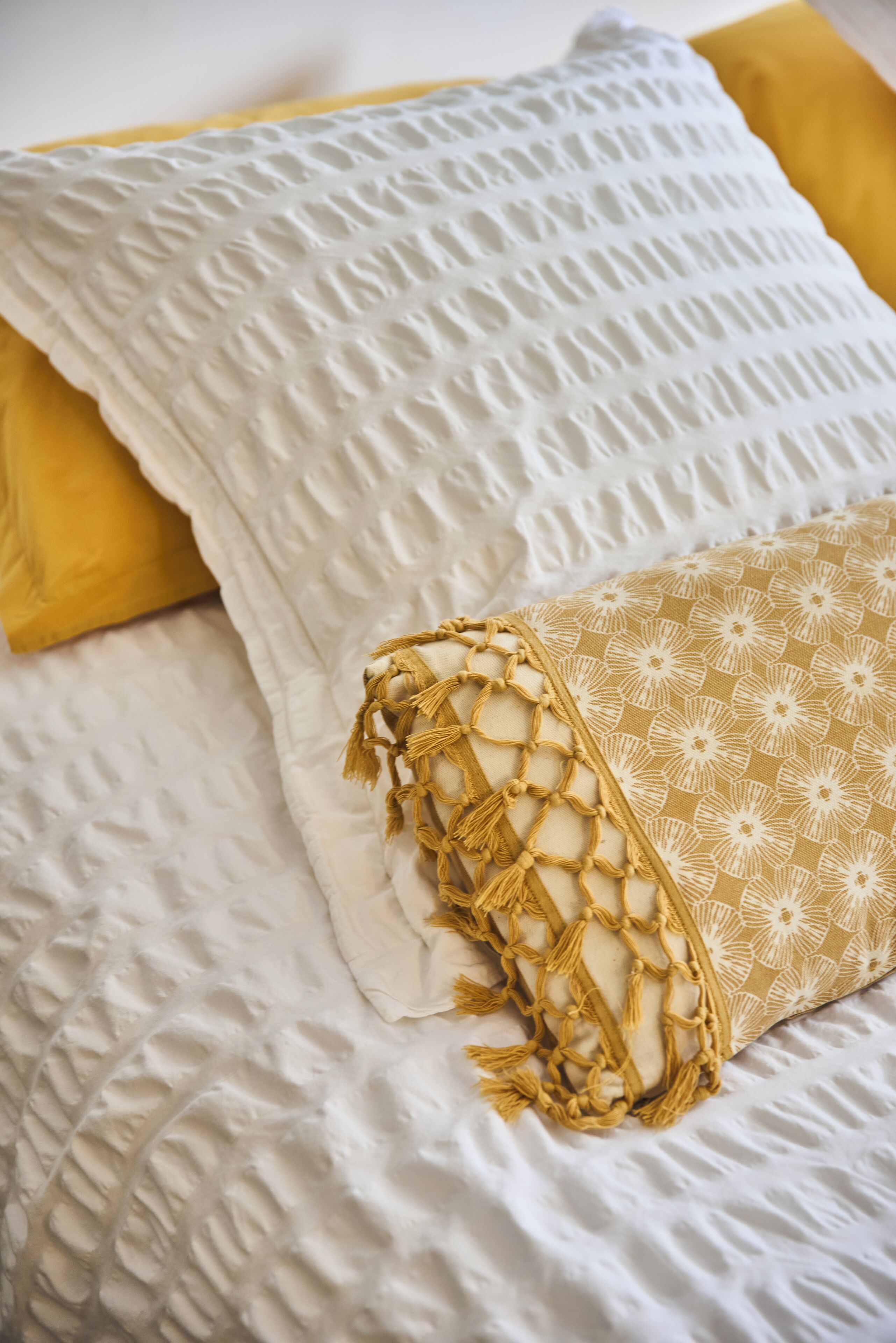 A bed with white and yellow pillows, a textured white blanket, and a patterned beige pillow.