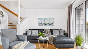 Sala de estar moderna com um sofá cinzento, uma poltrona cinza e uma mesa de centro de vidro. Há um quadro de veleiros na parede, uma escada com degraus de madeira e uma janela grande com cortinas.