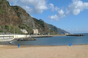 Una playa con un acantilado rocoso, un edificio con balcón con vistas al mar y un cielo despejado.