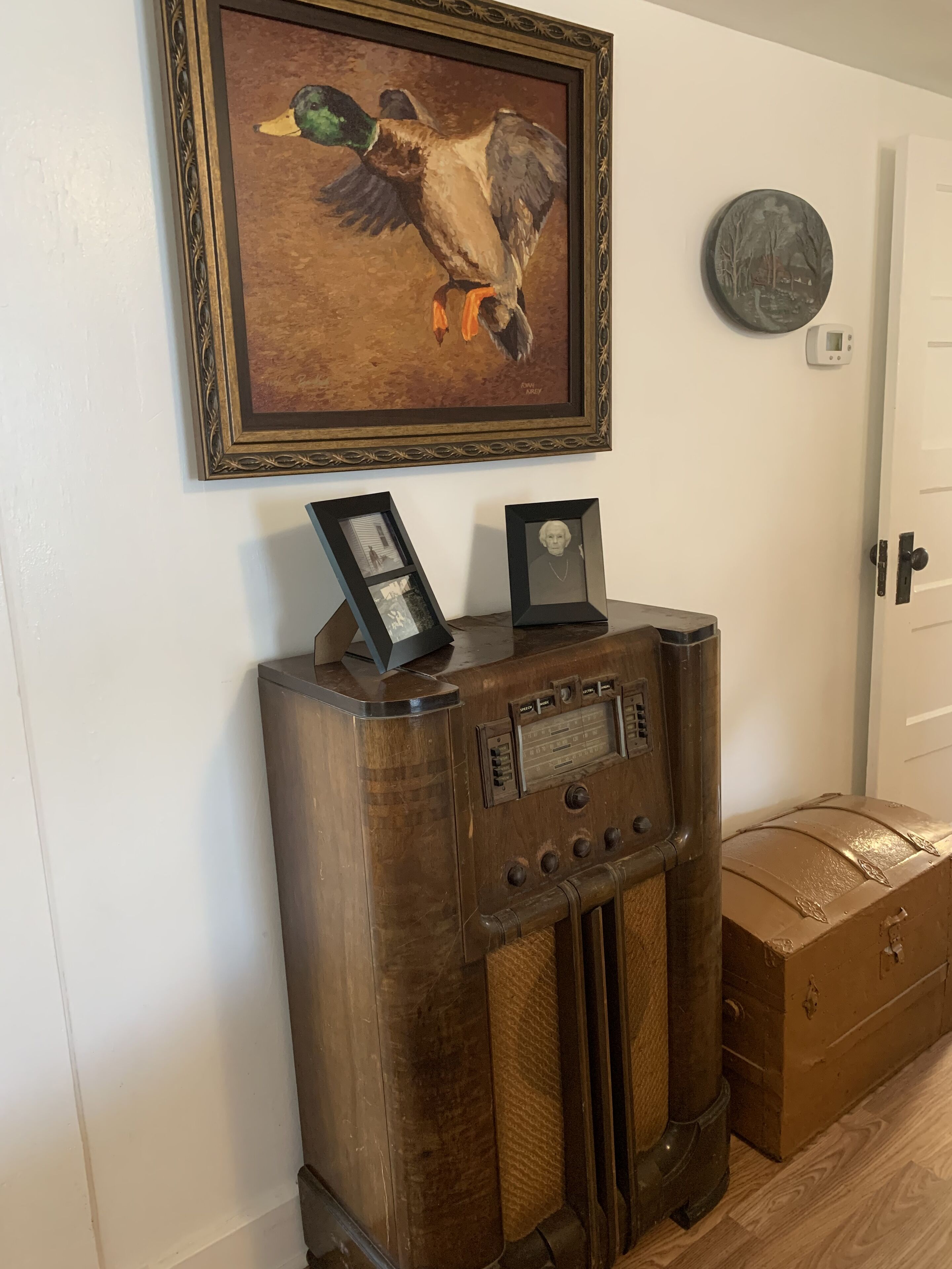 Vanha radio puukehikon päällä, kehystetty ankankuva ja termostaatti seinällä.