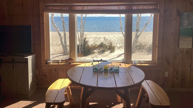 A wooden cabin with a large window overlooking a beach. Inside, there's a wooden table with benches and a sign above the window that reads "Dream of Summers That Last Forever."