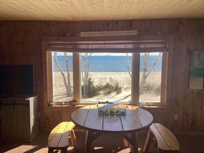 Une cabane en bois dotée d’une grande fenêtre donnant sur une plage. À l’intérieur, on trouve une table en bois avec des bancs, et au-dessus de la fenêtre, une pancarte sur laquelle on peut lire : « Rêvez d’étés qui ne finissent jamais. »