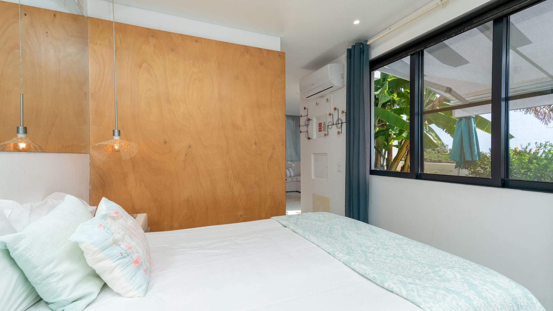 A bedroom with a wooden headboard, a bed with floral bedding, a hanging light fixture, and a window with a view of greenery.