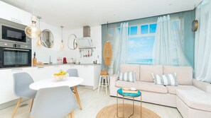 Una cocina moderna con horno empotrado, un comedor con mesa y sillas, un sofá con cojines y vistas al mar a través de una ventana con cortinas.