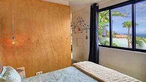 A bedroom with a large window, a bed with a patterned bedspread, a hanging light fixture, and a wooden wall.