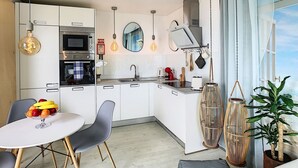 A modern kitchen with white cabinets, a built-in oven, a microwave, and a sink. There is a dining area with a table and chairs, and a large window with curtains.