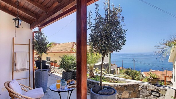 A covered patio with wicker chairs, a small table with breakfast items, potted plants, and a view of the sea. - Pôr do Sol by LovelyStay (Arco Da Calheta)