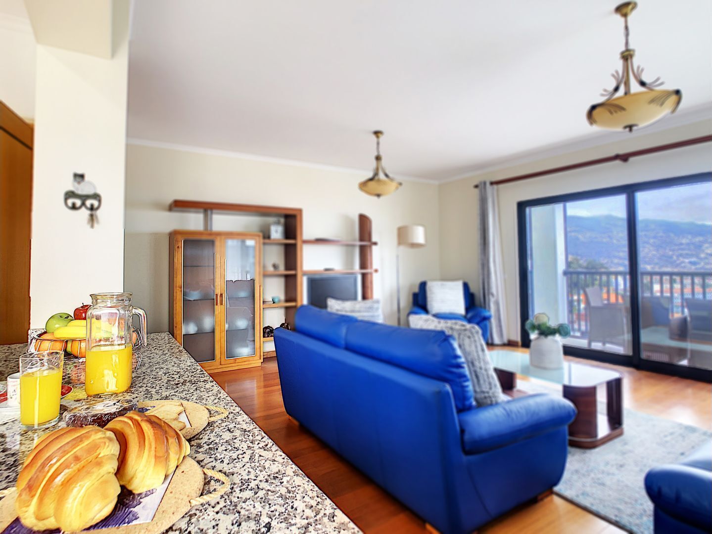 A modern living room with a blue sofa, a dining area with a granite countertop, and a large window offering a city view.