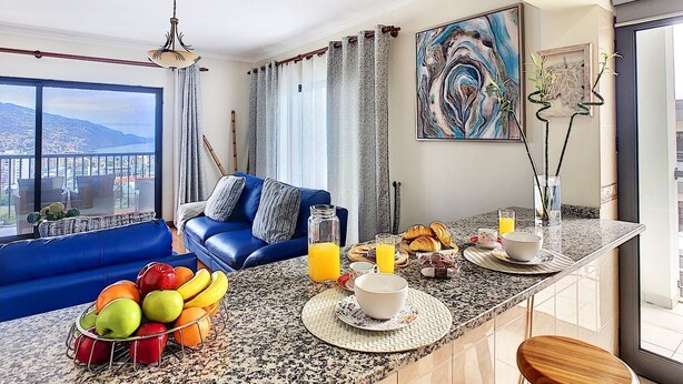 A modern kitchen with a blue sofa, breakfast setup, and a city view. - Gemini Apartment by LovelyStay (Funchal)
