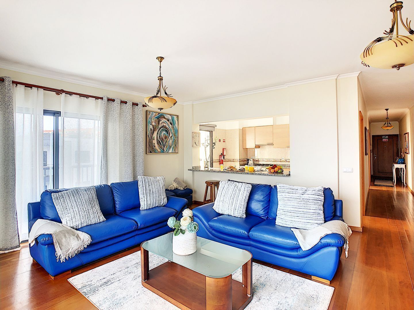 Una sala moderna con un conjunto de sofá azul, una mesa de centro de vidrio y una cocina visible a través de una puerta abierta.
