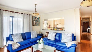 A modern living room with a blue sofa set, a glass coffee table, and a kitchen visible through an open door. - Gemini Apartment by LovelyStay (Funchal)
