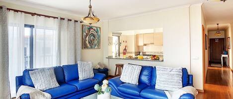 Una sala moderna con un conjunto de sofá azul, una mesa de centro de vidrio y una cocina visible a través de una puerta abierta.