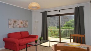 A modern living room with a red sofa, a glass coffee table, and a wooden chair. A large sliding glass door leads to a balcony with a view of greenery and a rocky landscape. - Apt. El Molino 2B  (Valle Gran Rey)