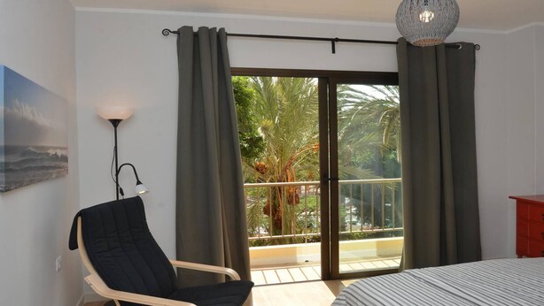 Een slaapkamer met een bed, een stoel, een nachtkastje, een lamp, een balkon met uitzicht op palmbomen en een schilderij aan de muur.