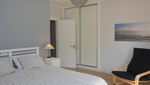 Een slaapkamer met een bed, nachtkastje, stoel, garderobe en een schilderij van de oceaan.
