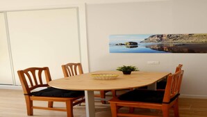 Een eethoek met een houten tafel en stoelen, een kleine potplant en een schilderij van een kustlandschap aan de muur.