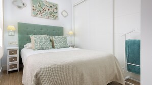 A bedroom with a green upholstered headboard, two bedside tables with lamps, a bed with patterned pillows, a wall-mounted artwork, and a towel rack with a towel. - Genteel Home Juan Rabadan I (Seville)