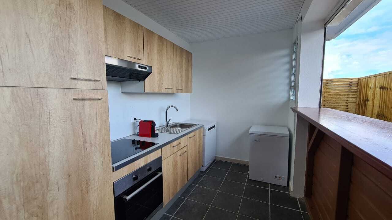 Ett kompakt kök med träskåp, en svart bänkskiva, en röd vattenkokare, ett handfat, ett litet vitt kylskåp och ett fönster med utsikt över en träveranda.