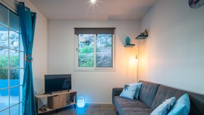 Ein Wohnzimmer mit Fernseher auf Holzfuß, ein graues Sofa mit gemusterten Kissen, einer Stehlampe und einem Fenster mit Vorhängen.