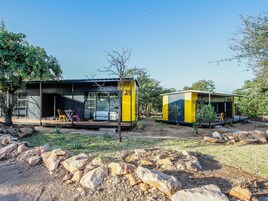 Duas cabanas amarelas e pretas com área de estar externa, cercadas por pedras e vegetação rasteira.