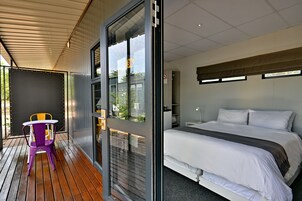 Quarto de hotel moderno com uma cama grande, uma mesa pequena com cadeiras e uma porta de vidro deslizante para uma área externa.