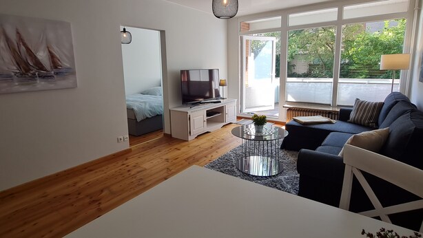 A modern living room with a sofa, a TV, and a dining table. There is a large window providing a view of greenery outside. - Modern city apartment with balcony and private parking space in a quiet location (Kiel)