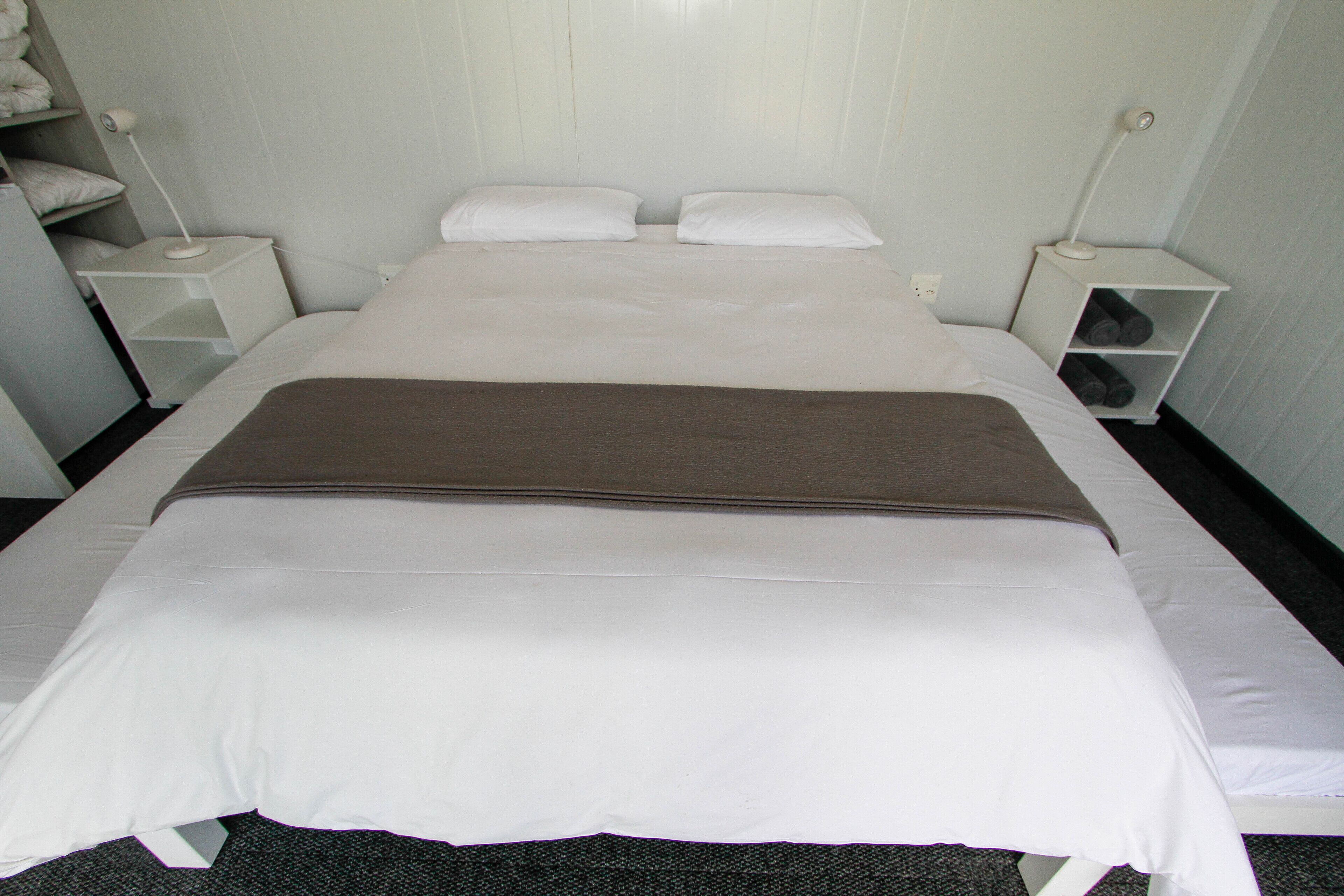 A neatly made bed with white linens and a brown blanket, flanked by white nightstands with lamps, in a room with light-colored walls.