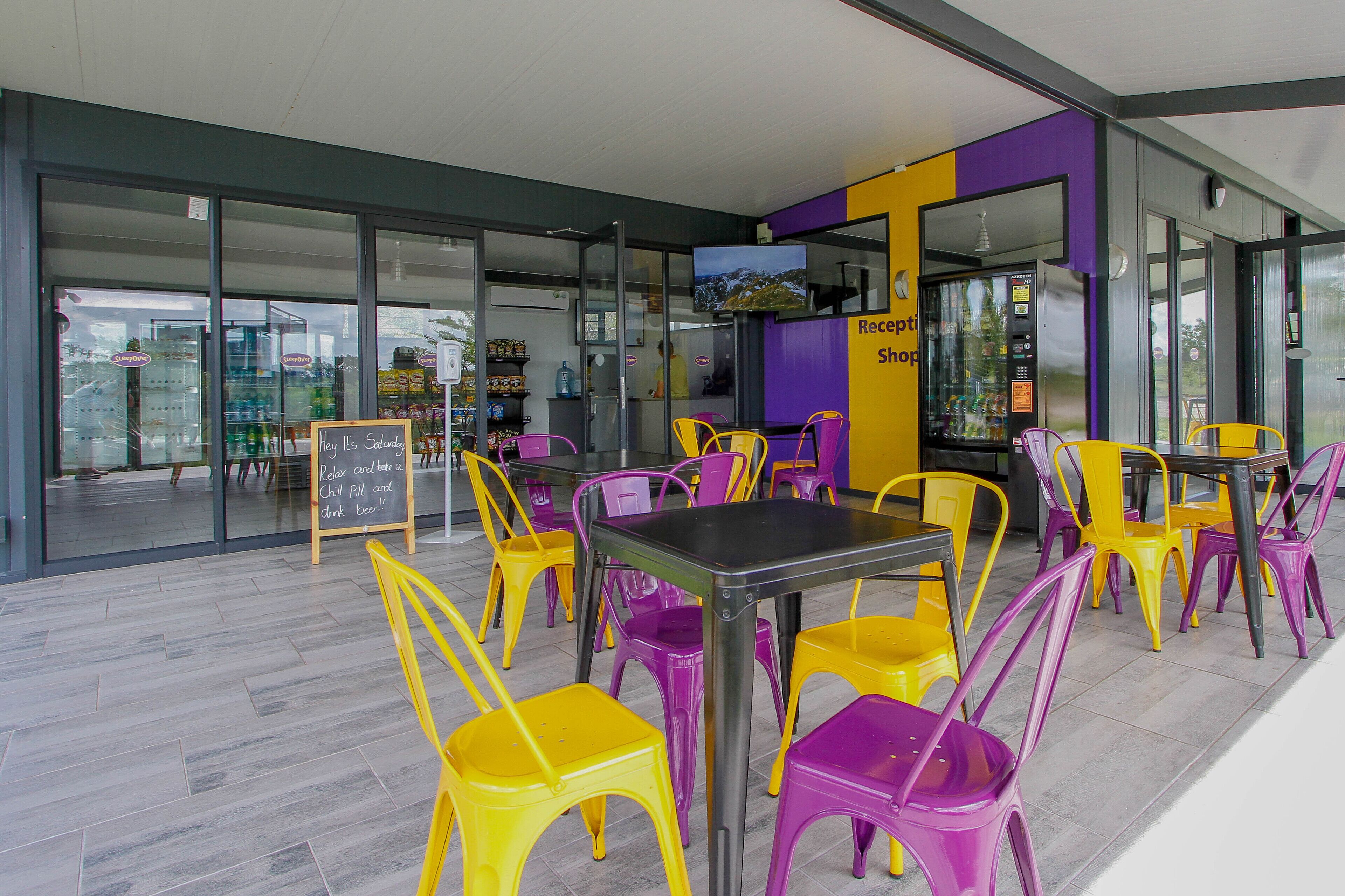 An outdoor seating area with yellow and purple chairs, a black table, and a sign that reads "Relax and enjoy a chill pill and drink beer."