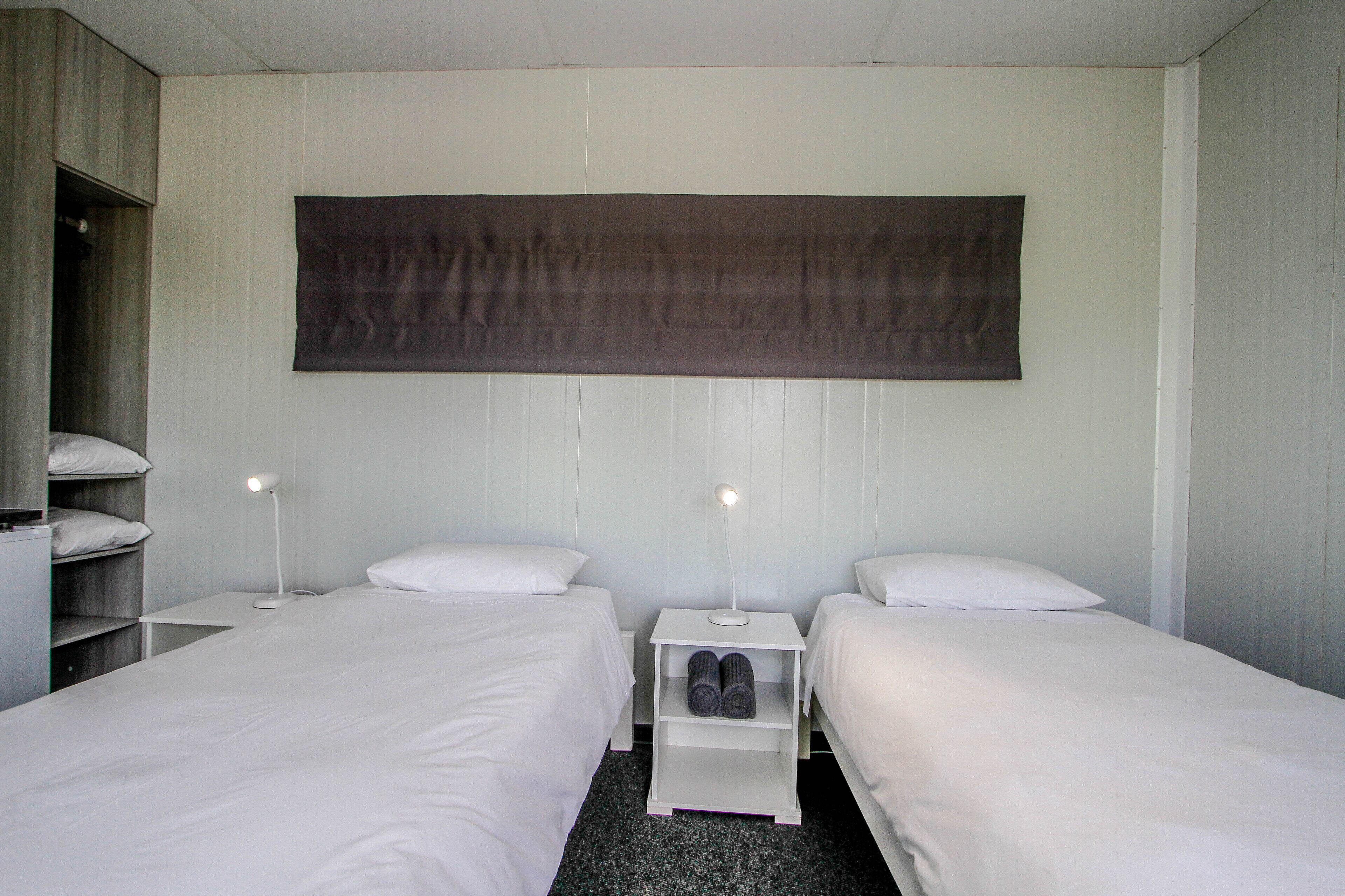 A room with two single beds, a large dark curtain, a white nightstand with a lamp, and a shelf with rolled towels.