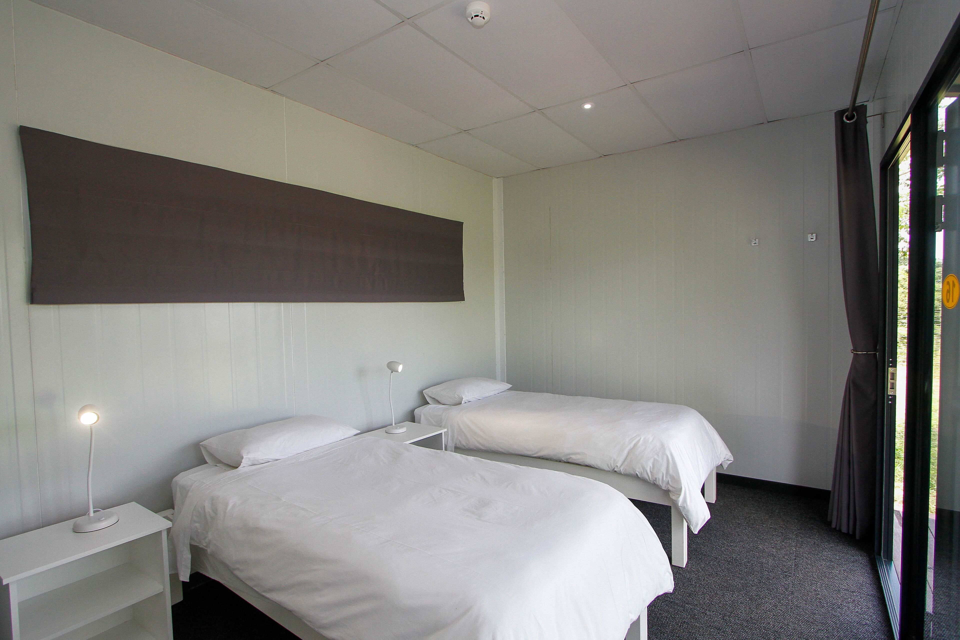 A hotel room with two single beds, a bedside table with a lamp, and a large window with curtains.