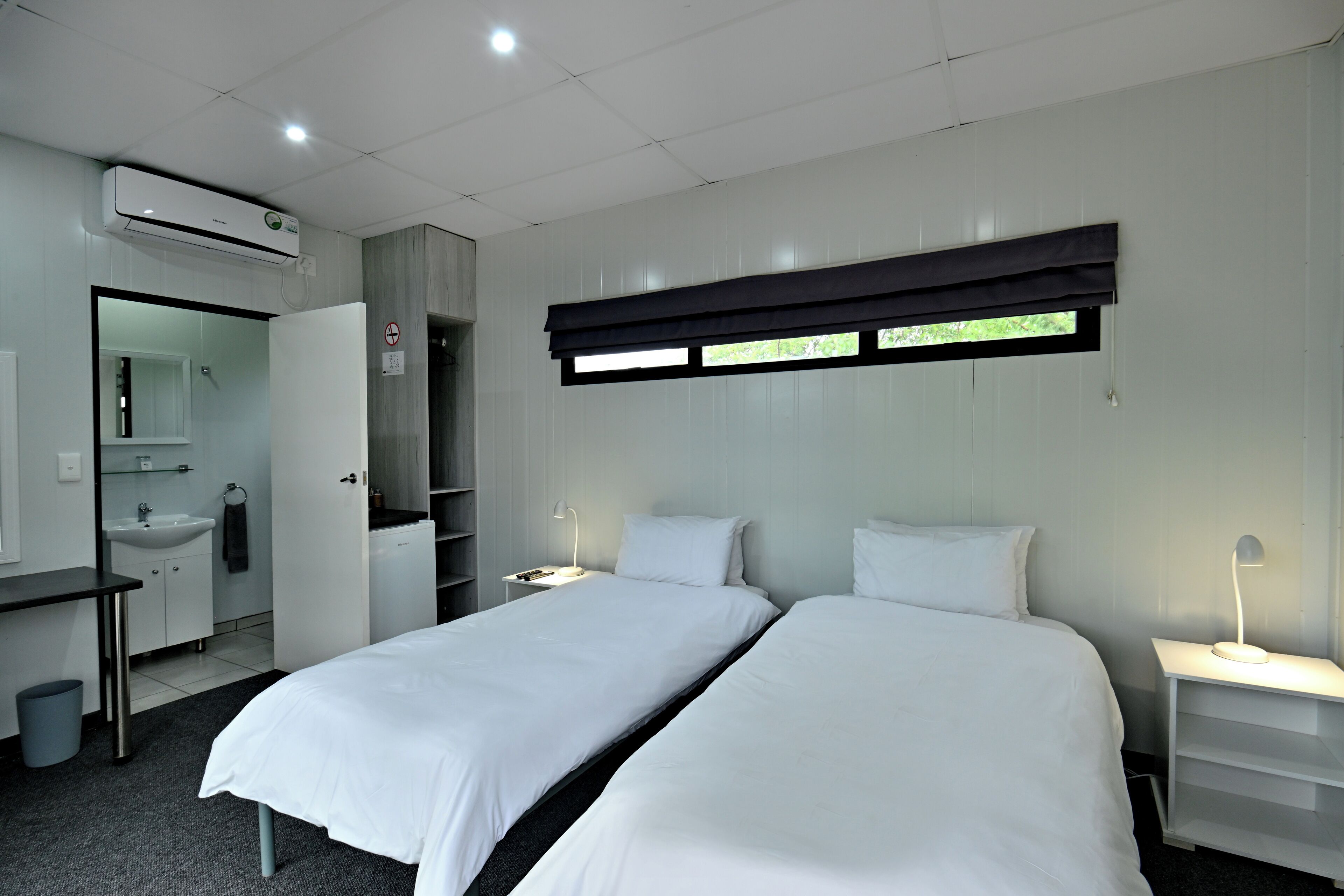 Een hotelkamer met twee eenpersoonsbedden, een bureau met stoel, een kaptafel met spiegel, een wastafel en een airconditioning.