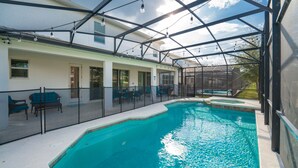 A covered outdoor swimming pool with a glass enclosure, seating area, and string lights.