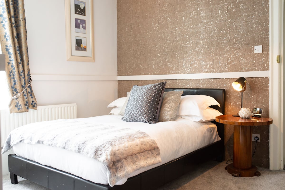 A neatly made bed with a white and gray blanket, a round wooden nightstand with a lamp, and a wall with a textured finish.