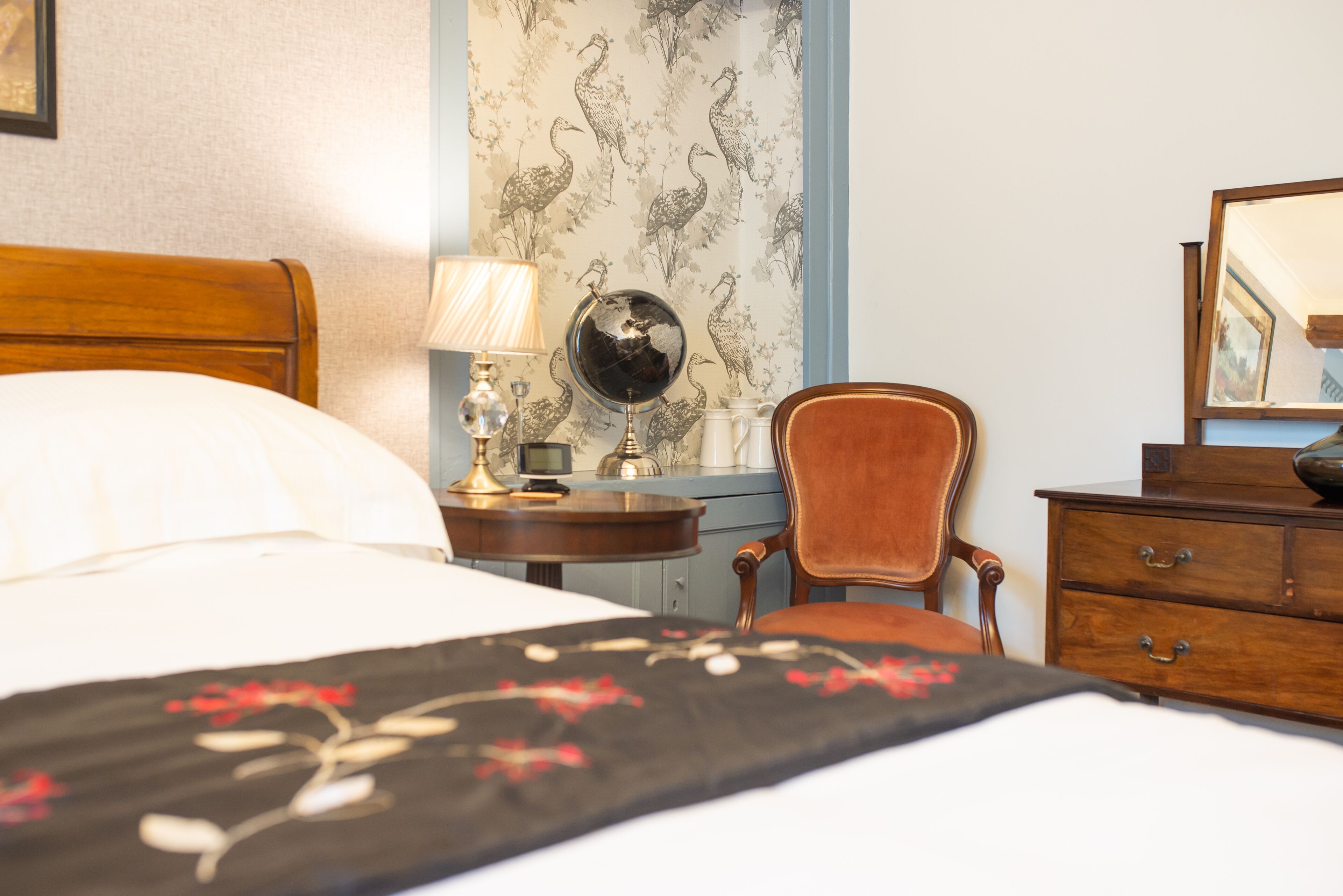 A neatly arranged hotel room with a bed, a chair, a dresser, and a nightstand with a lamp and a globe.