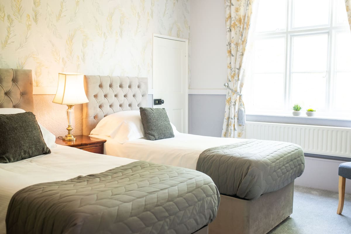 A hotel room with two beds, a nightstand with a lamp, a window with curtains, and a radiator.
