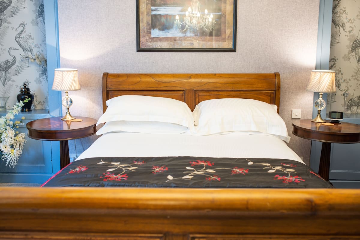 A neatly made bed with a floral bedspread, two bedside tables with lamps, a framed picture on the wall, and floral wallpaper.