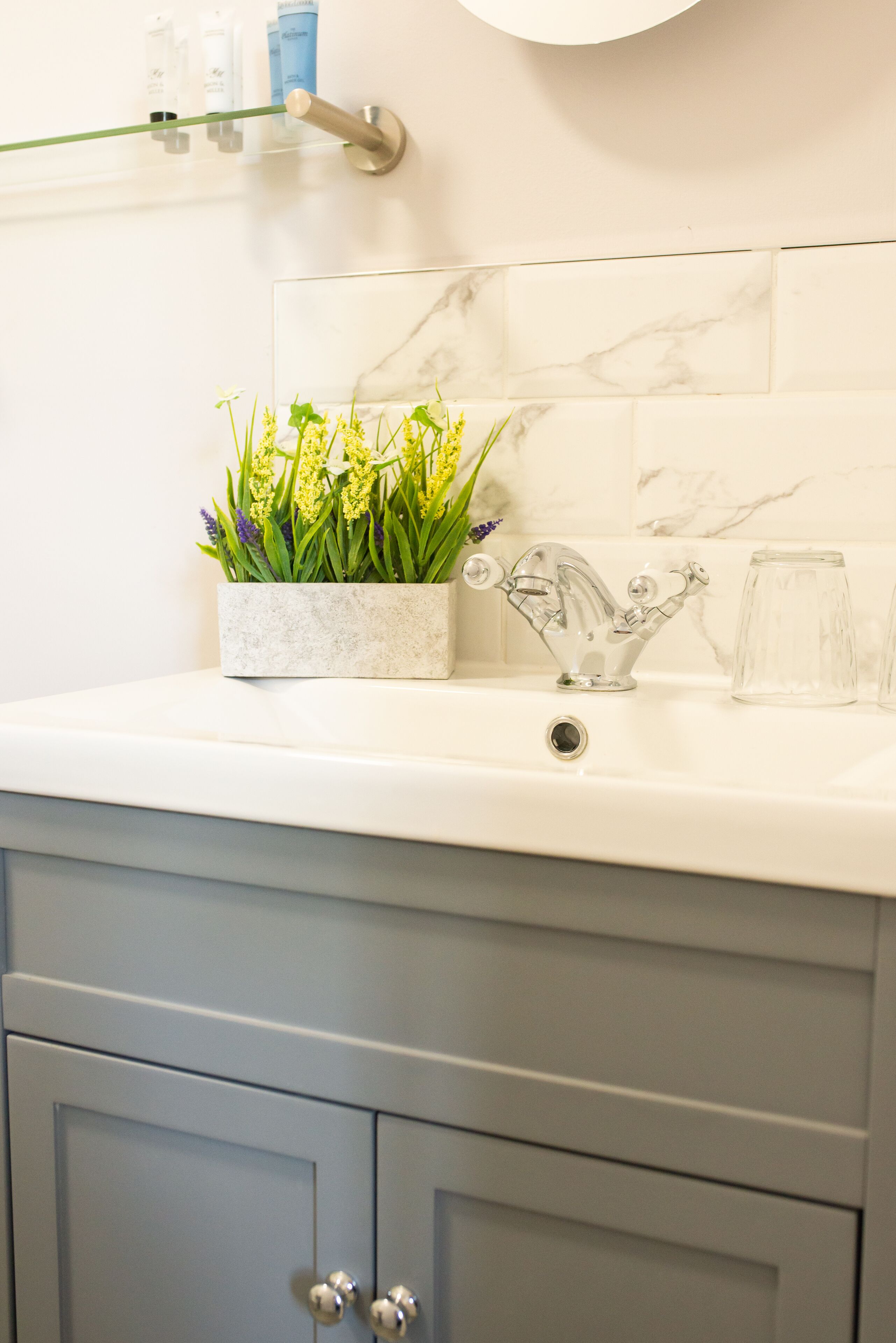 Une salle de bain avec un pare-baignoire en marbre, un lavabo blanc, un vase en verre avec des fleurs et une carafe en verre.