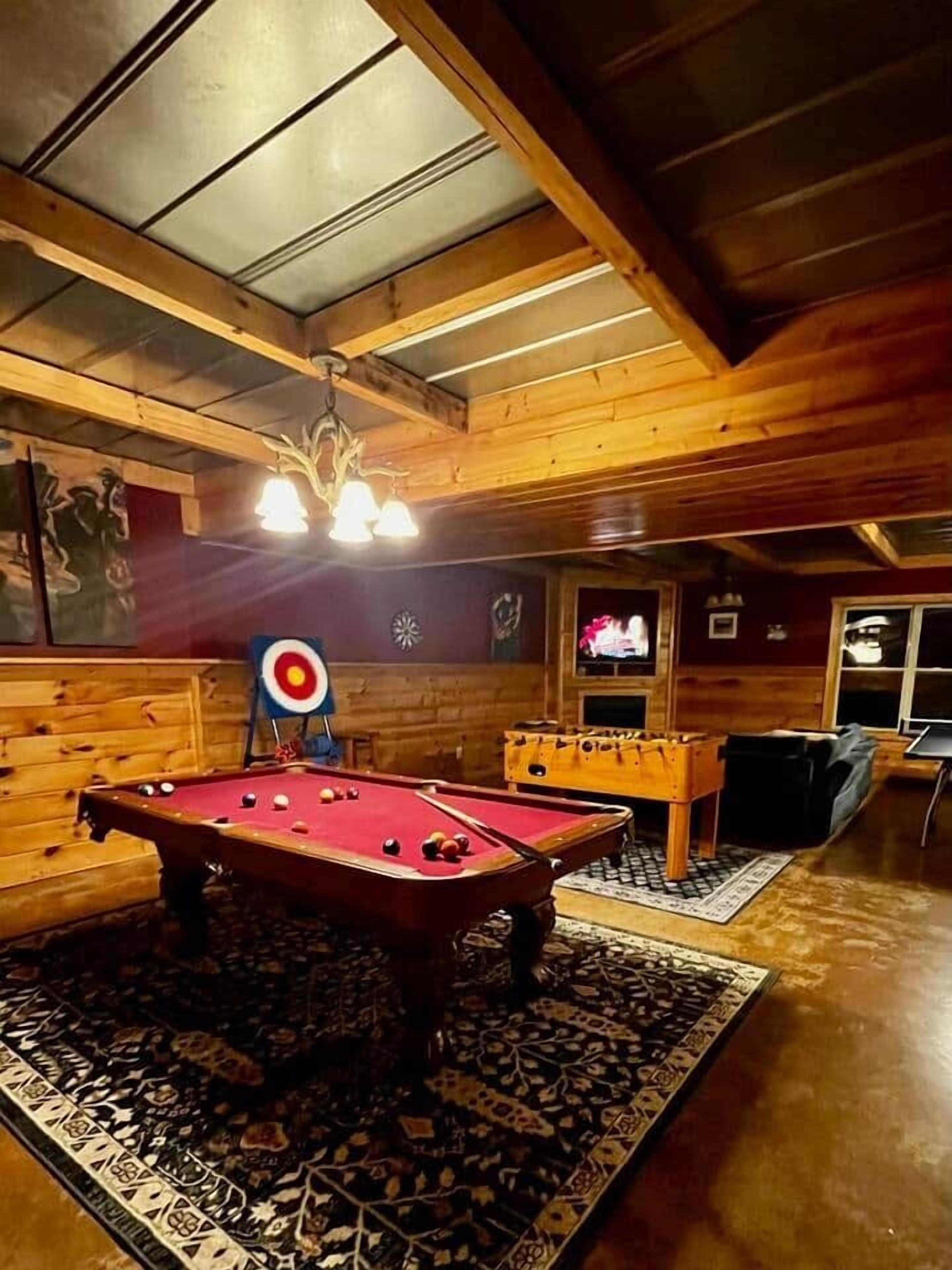 A pool table in a wooden room with a target on the wall, a foosball table, and a television.
