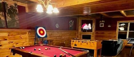 A pool table in a wooden room with a target on the wall, a foosball table, and a television.