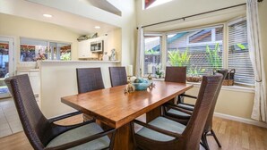 Een eethoek met een houten tafel en rieten stoelen, een keuken op de achtergrond, en een raam met zonwering.