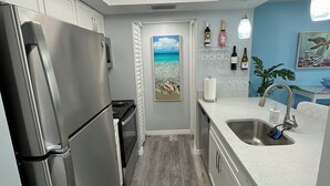 Een moderne keuken met een roestvrijstalen koelkast, een inbouwoven en een spoelbak met een kraan. Er is een plafondlamp en een deur met een schilderij van een zandstrand.