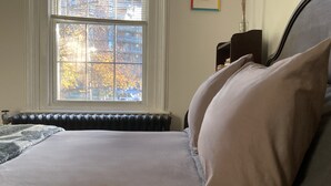 A bedroom with a large window, a radiator, a bed with pillows, a framed picture on the wall, and a vase with dried flowers. - Spacious Egleston Sq 3 Bedroom, Dog Friendly (Boston)