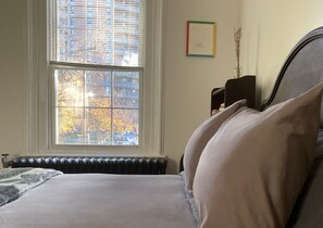 A bedroom with a large window, a radiator, a bed with pillows, a framed picture on the wall, and a vase with dried flowers. - Spacious Egleston Sq 3 Bedroom, Dog Friendly (Boston)