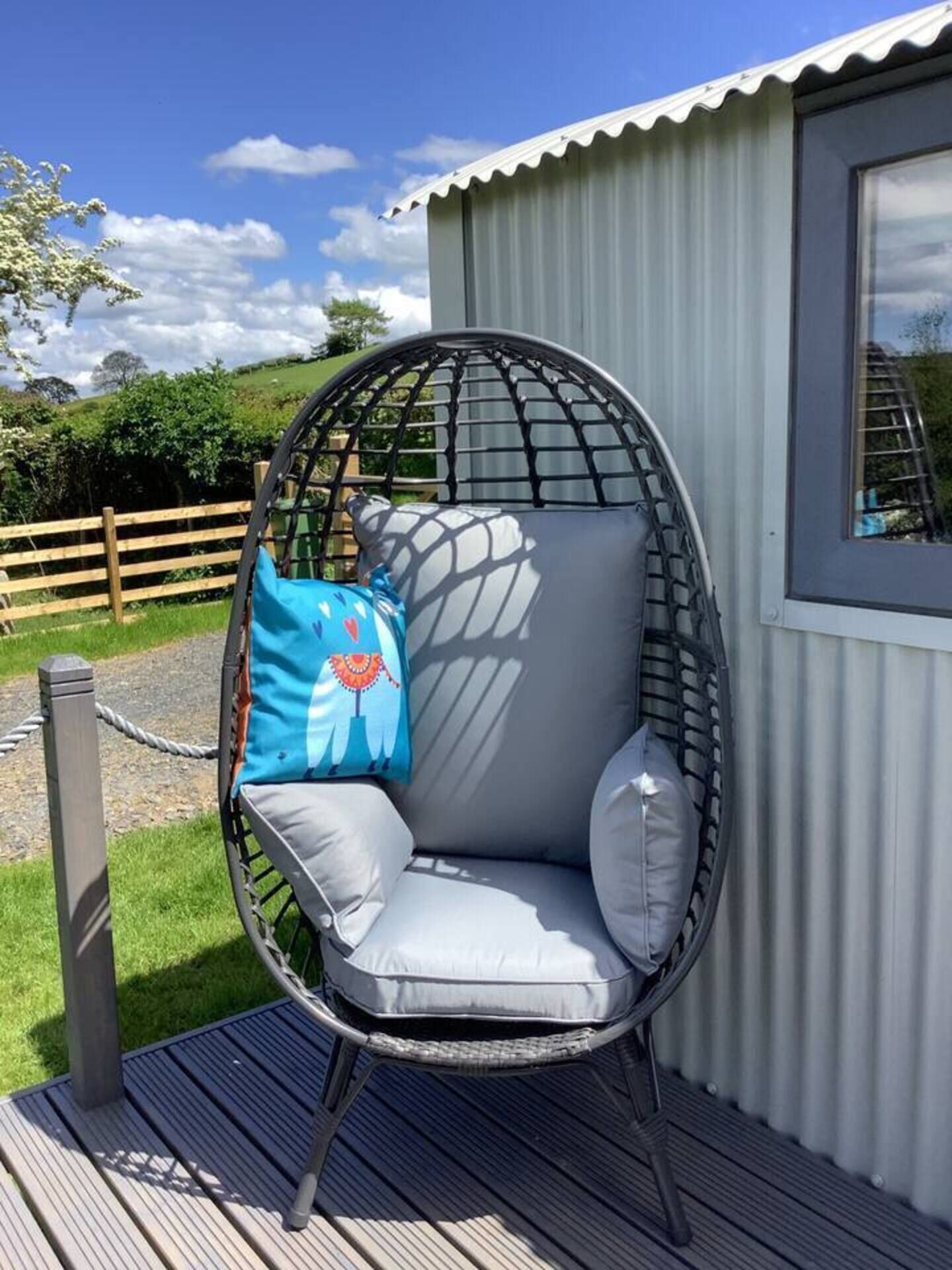 A round, wire-framed chair with grey cushions, positioned on a wooden deck next to a corrugated metal building.