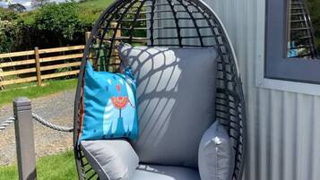 A round, wire-framed chair with grey cushions, positioned on a wooden deck next to a corrugated metal building.