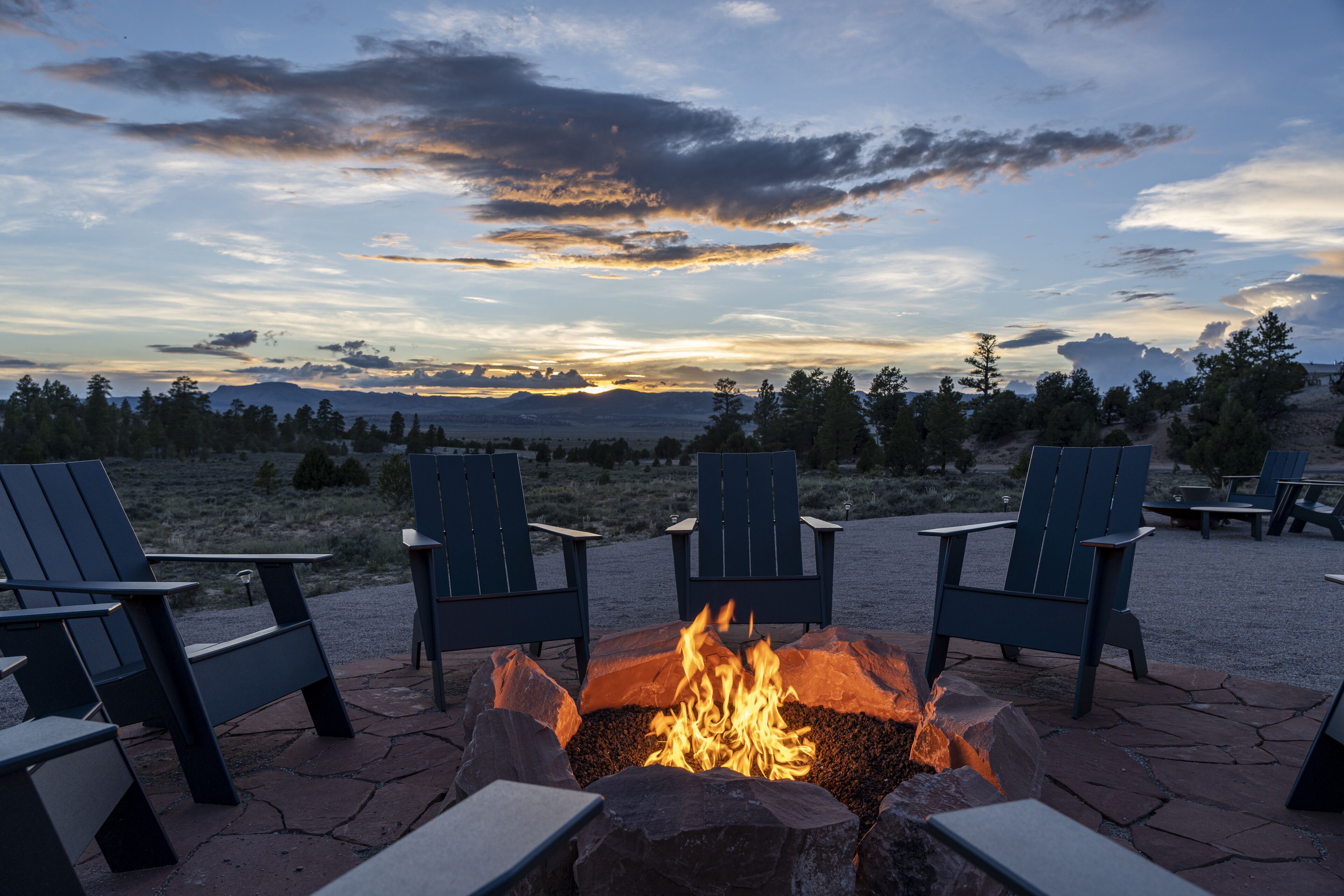 A fire pit with chairs around it, set on a stone patio with a view of trees and mountains in the background.