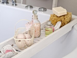 A bathroom vanity with a white sink, a glass soap dispenser, a bottle of liquid soap, a sponge, and a basket with bath items.