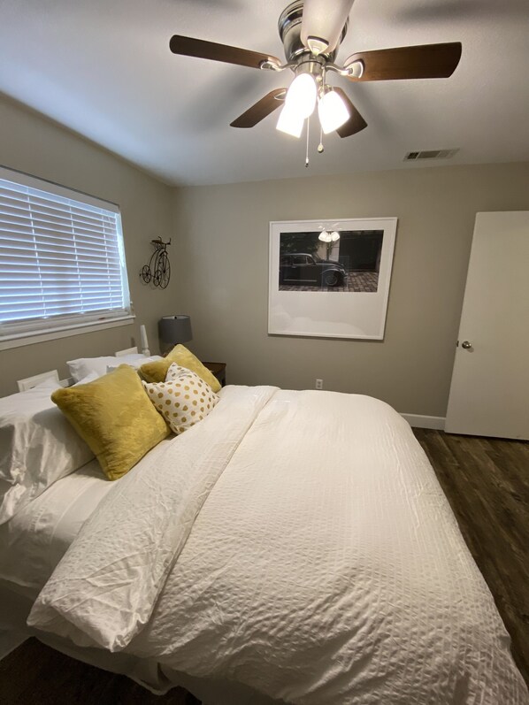 A bedroom with a bed, a ceiling fan, a window with blinds, a framed picture on the wall, and a door.