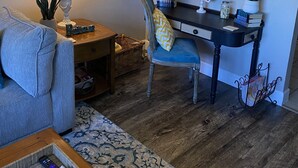 A cozy living room with a blue armchair, a wooden coffee table, a desk with a chair, a clock, and a mirror. - The View at 5 Hills (Copperas Cove)