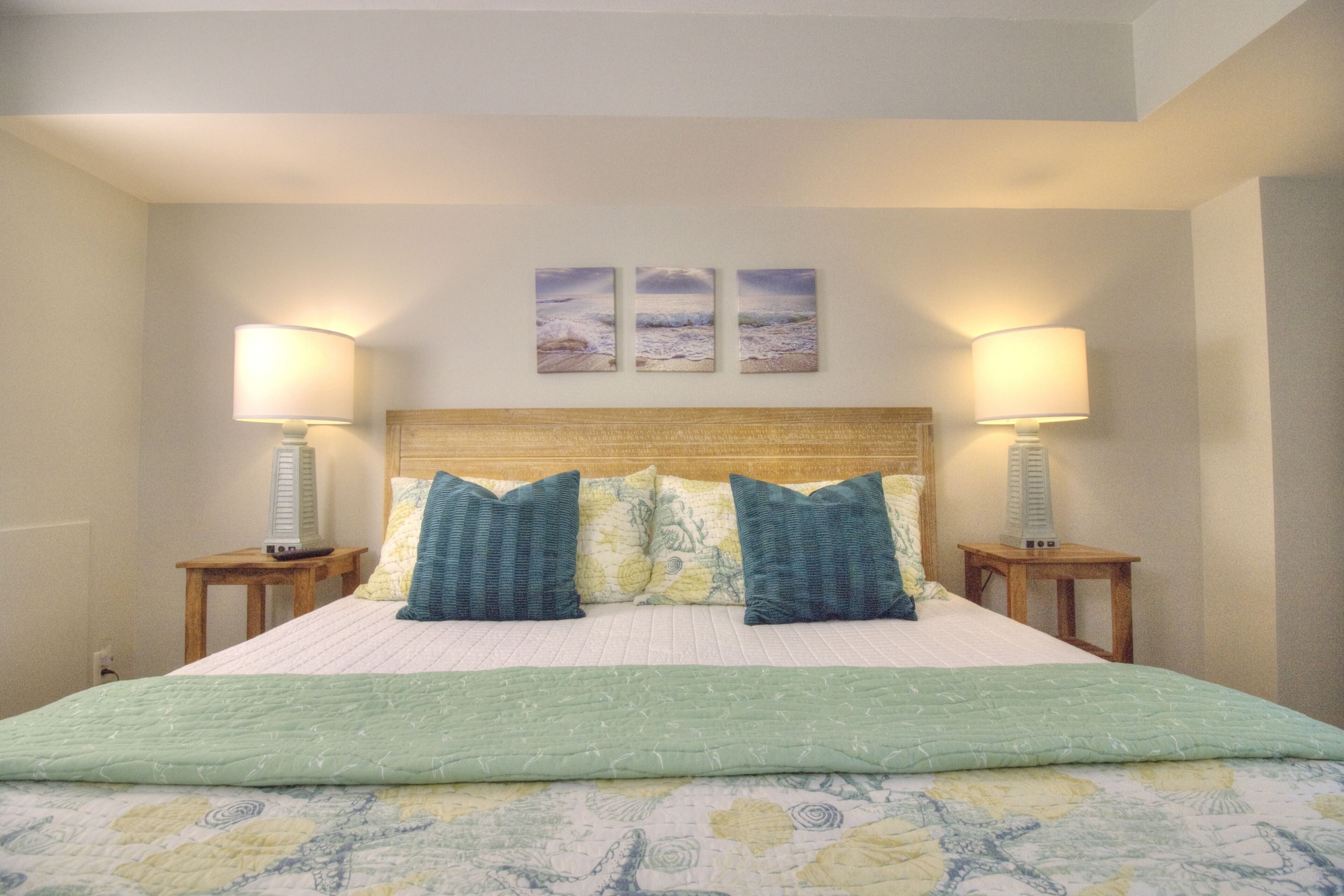A neatly made bed with a green quilt, two blue pillows, and a headboard with a wooden panel. There are two bedside lamps on wooden nightstands, and three framed pictures on the wall above the bed.